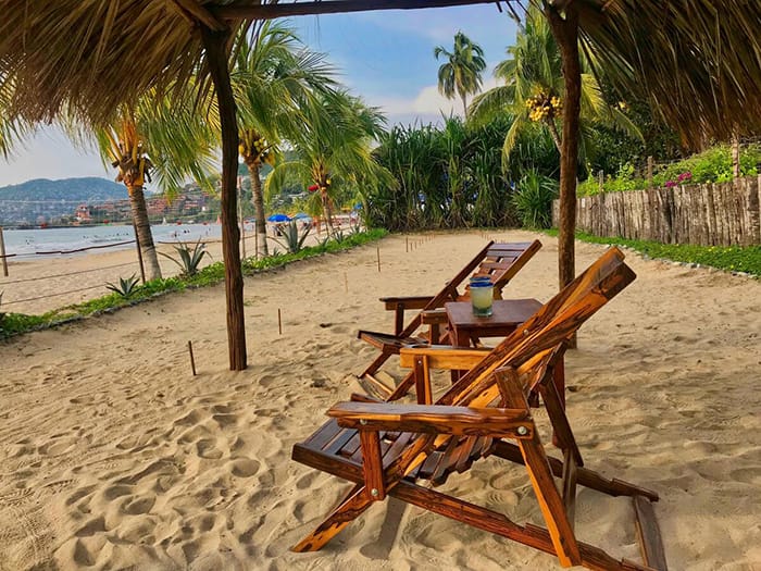 Zona de playa de descanso con dos sillas de madera y palmeras en Casa de los pájaros en Airbnb en México