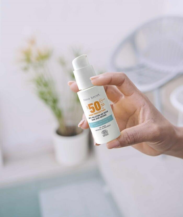 Mano sujetando un bote de protección solar de Alma Secret y de fondo la piscina con una maceta