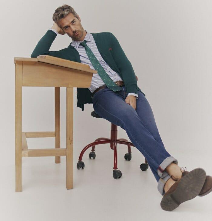 Hombre sentado en una silla de ruedas y apoyado en una mesa de madera con vaqueros, camisa, corbata y chaqueta de Scotta