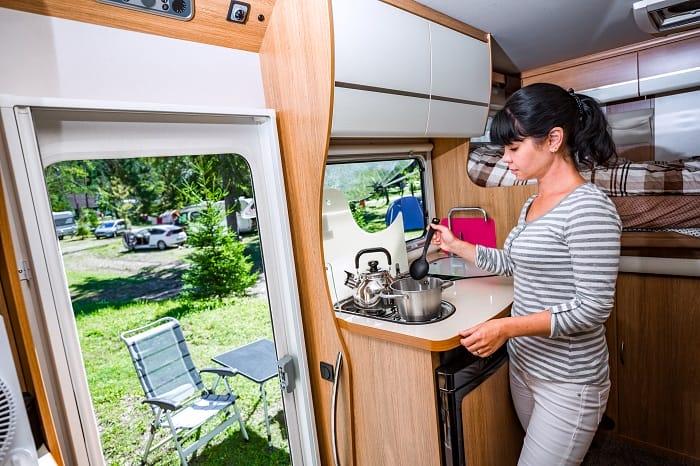 Mujer cocinando dentro de su camper con la puerta abierta y de fondo el campo