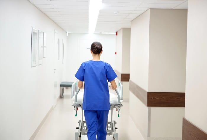 Cómo trabajar en un hospital público. Mujer sanitaria de espaldas llevando una camilla por los pasillos de un hospital