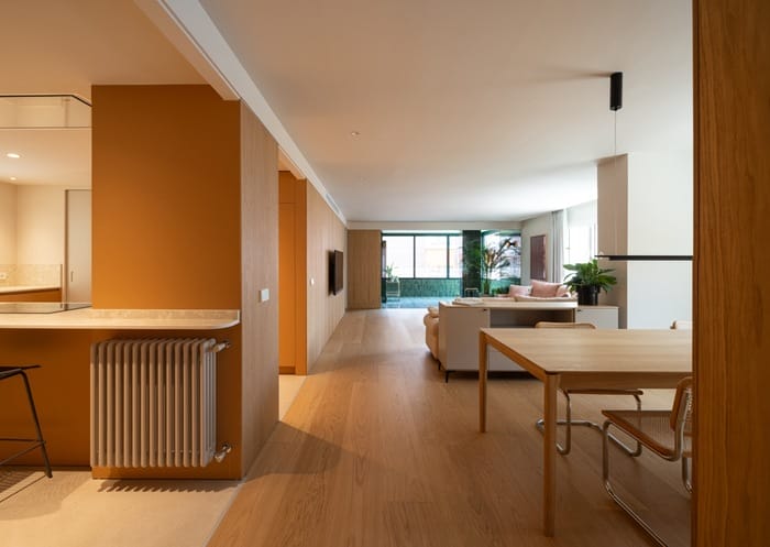 Perspectiva abierta entre cocina, comedor y salón con tonos cálidos y pavimento continuo de madera