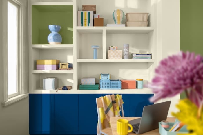 Estantería moderna en tonos blancos con objetos decorativos y cajas de colores, combinada con muebles inferiores en azul intenso y pared verde, iluminada por luz natural en un espacio de trabajo contemporáneo.