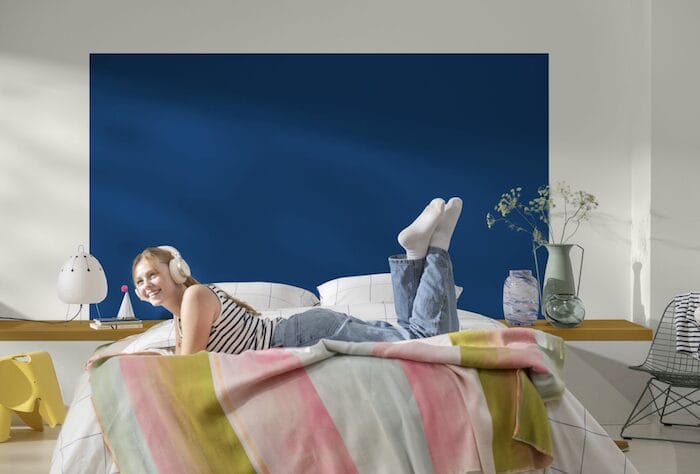 color del año de bruguer Dormitorio juvenil con mural azul intenso detrás de la cama y una joven disfrutando con auriculares sobre una colcha a rayas de colores suaves.