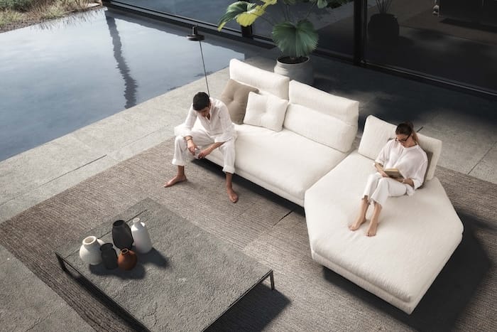 Sofá modular blanco con chaise longue en un espacio exterior junto a una piscina, con dos personas relajándose sobre alfombras neutras y decoración de jarrones.