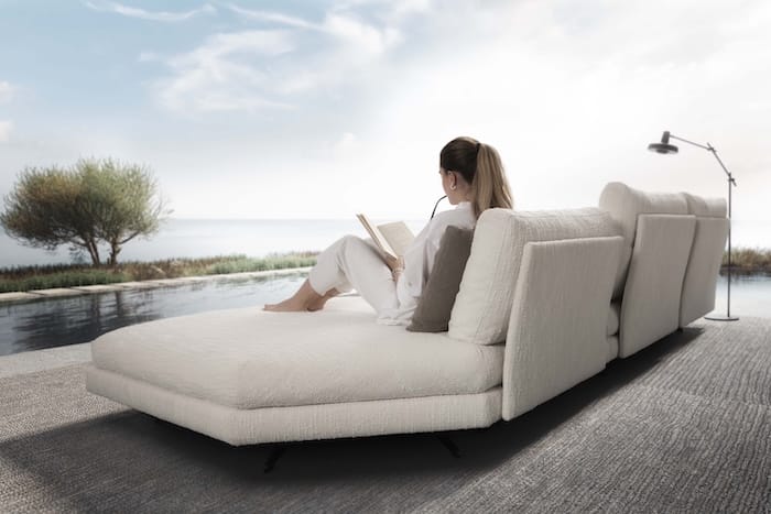 Chaise longue blanca con una mujer leyendo frente a una piscina y un paisaje natural junto al mar, con luz suave y ambiente relajante.